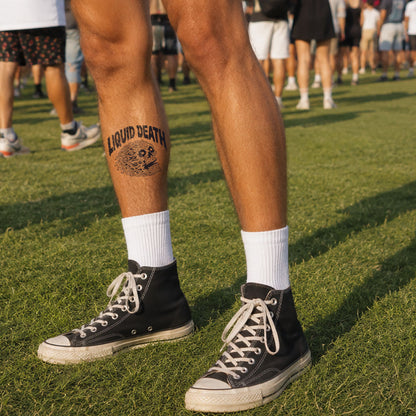 Person wearing black high-top sneakers with white laces on a grassy field, showing a tattoo on their leg.