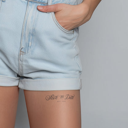 Person wearing light blue denim shorts with a 'Save the Date' tattoo on the leg against a gray background