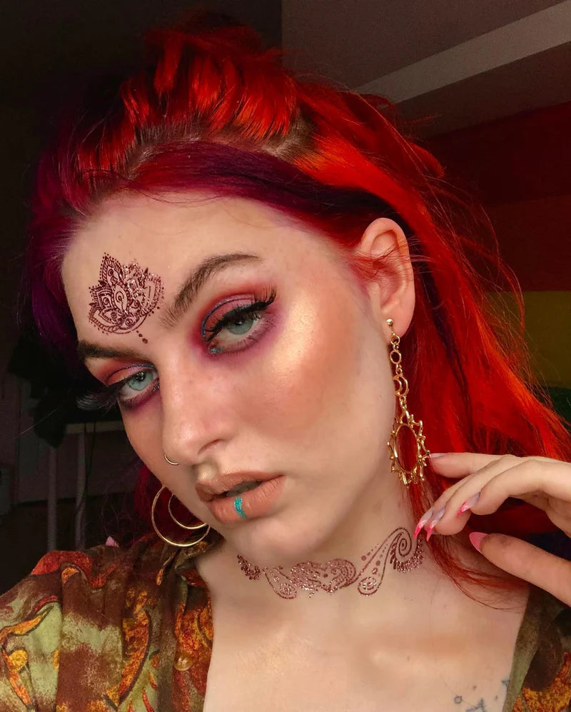 Person with red hair and intricate tattoos on face and neck, wearing gold earrings.
