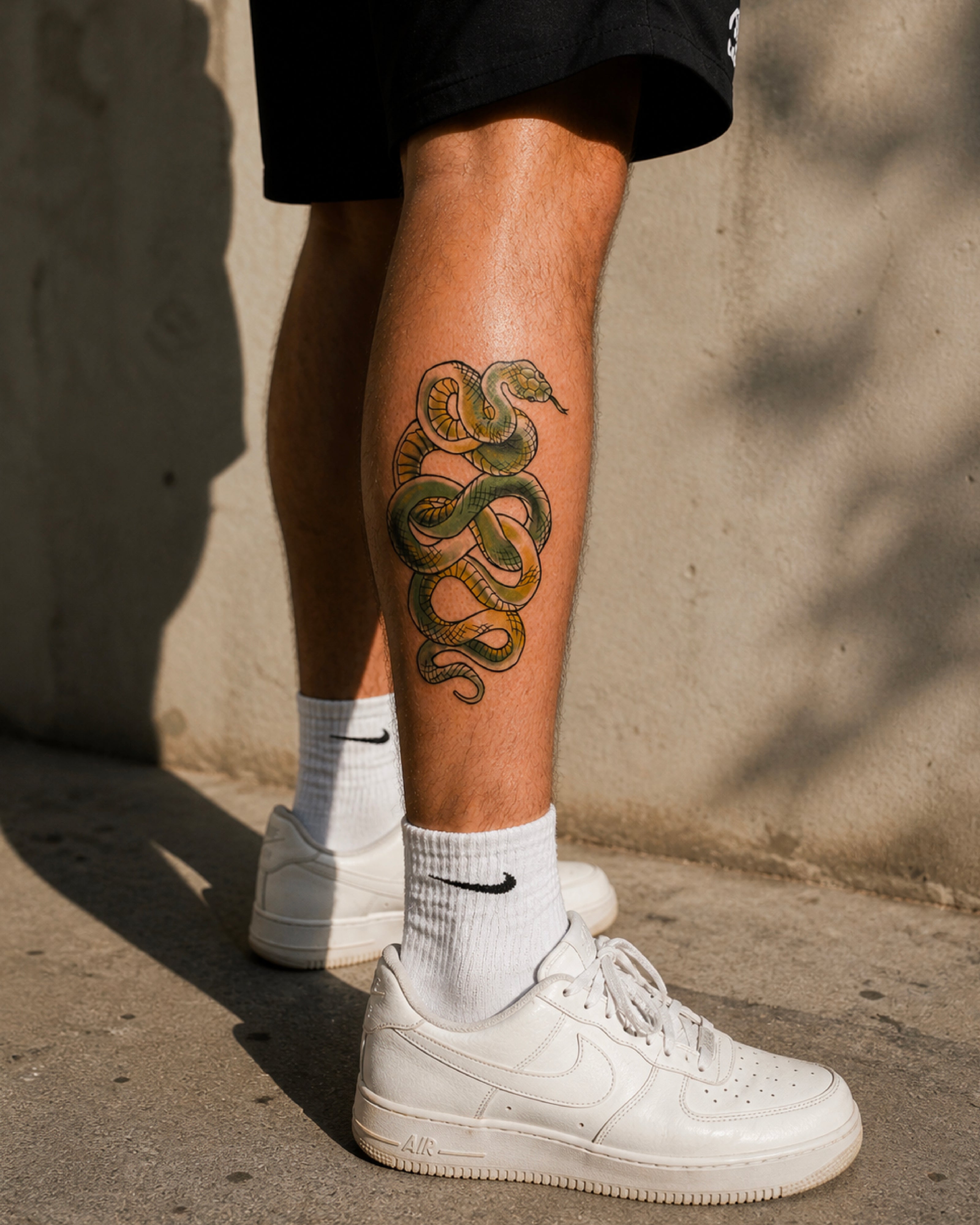 Person wearing white sneakers and socks with a snake tattoo on their leg against a concrete wall.