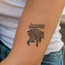 A temporary tattoo on a person's forearm featuring a raccoon and the name 'JIMOTHY' in stylized text, worn over a white ribbed top and blue jeans.