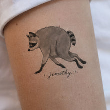 A temporary tattoo of a running raccoon with the name 'jimothy' written below in cursive, shown applied to a person's upper arm.
