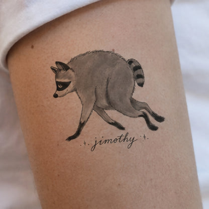 A temporary tattoo of a running raccoon with the name 'jimothy' written below in cursive, shown applied to a person's upper arm.