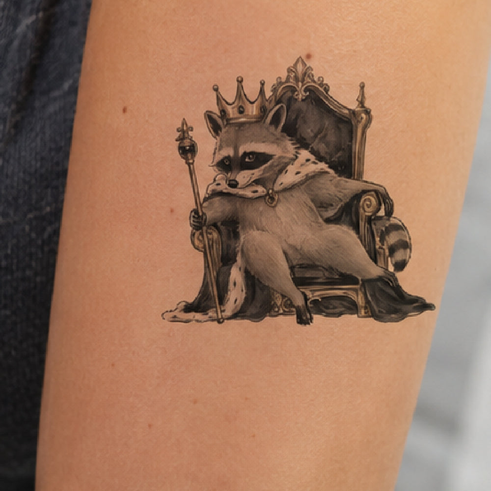 A temporary tattoo of a raccoon wearing a crown and sitting on an ornate throne, shown applied to a person's forearm.