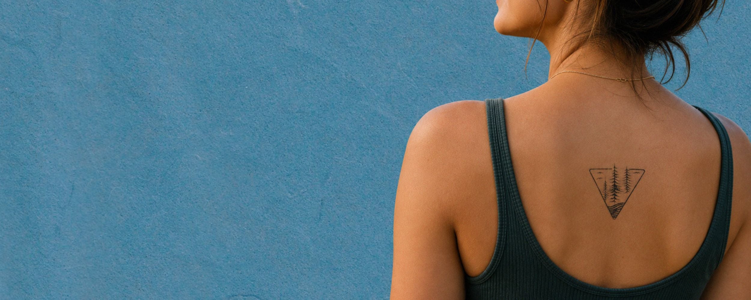 A woman shown from the back wearing a dark green ribbed tank top, displaying a small black geometric triangle tattoo on her upper back, set against a textured blue wall.