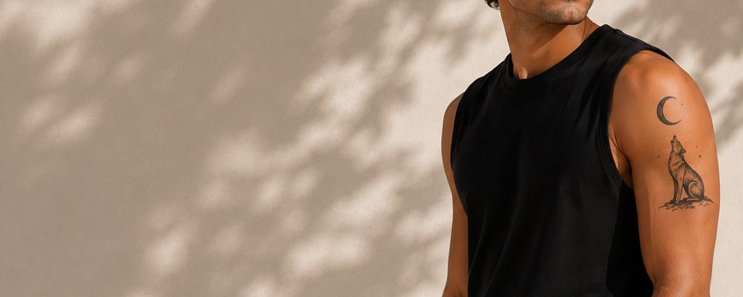 A man wearing a black sleeveless tank top, shown from the chest up against a beige wall with leaf shadows. The man has a small black line-art tattoo of a howling wolf and a crescent moon on his upper arm.